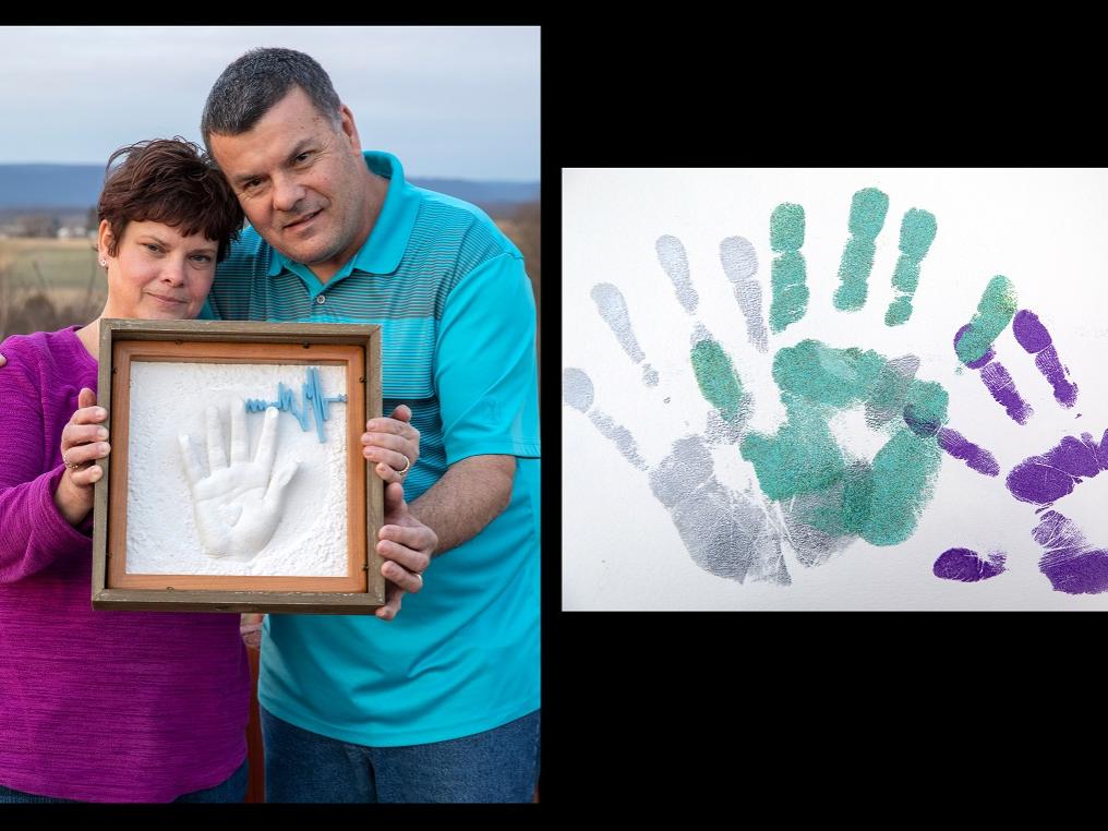 Wendy Gilbert, who has short hair and wears a bright top and jean, and Ronald Gilbert, who wears a polo shirt and jeans, hold a framed shadow box containing a resin handprint and 3D heartbeat. They are standing outside with a field in the background. Dylan’s handprints are beside them.