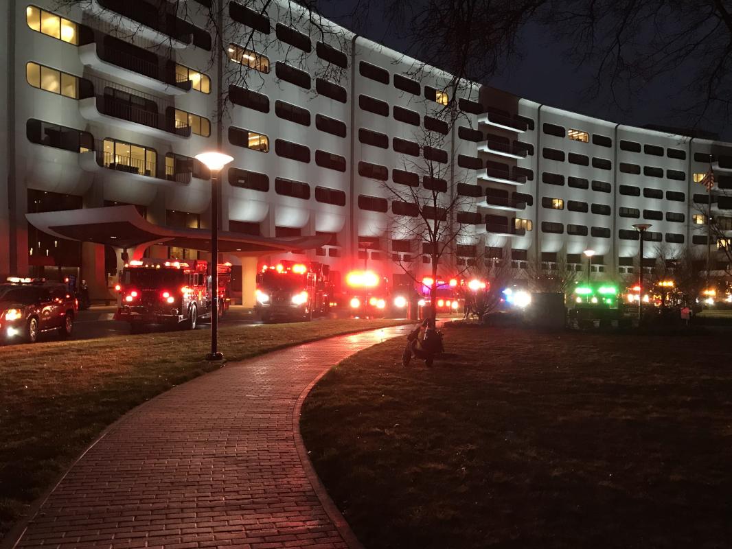Several emergency vehicles and tow trucks with flashing lights park along the crescent at Hershey Medical Center, at dusk.