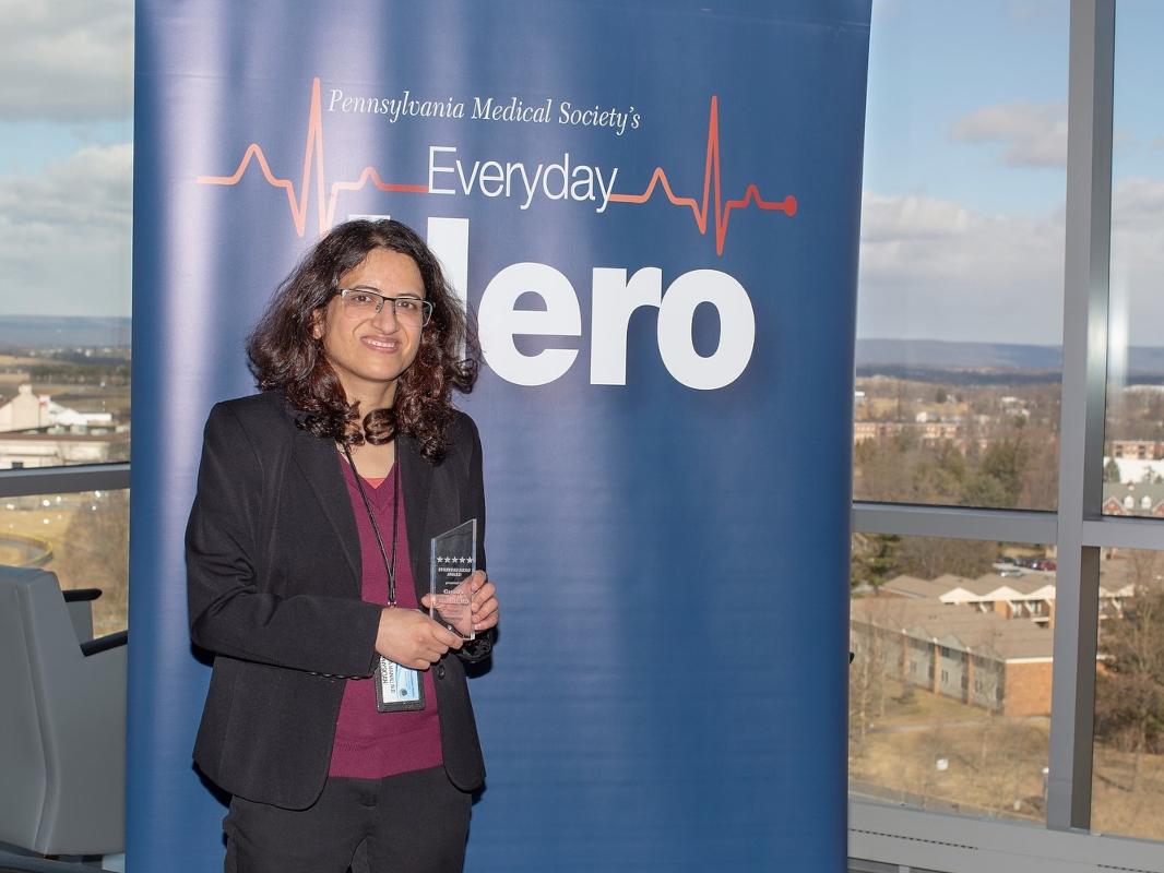 Dr. Gayatra Mainali, wearing professional attire, stands in front of a large pull-up banner with the words “Pennsylvania Medical Society’s Everyday Hero.” Buildings, trees and mountains are in the background, outside a window.