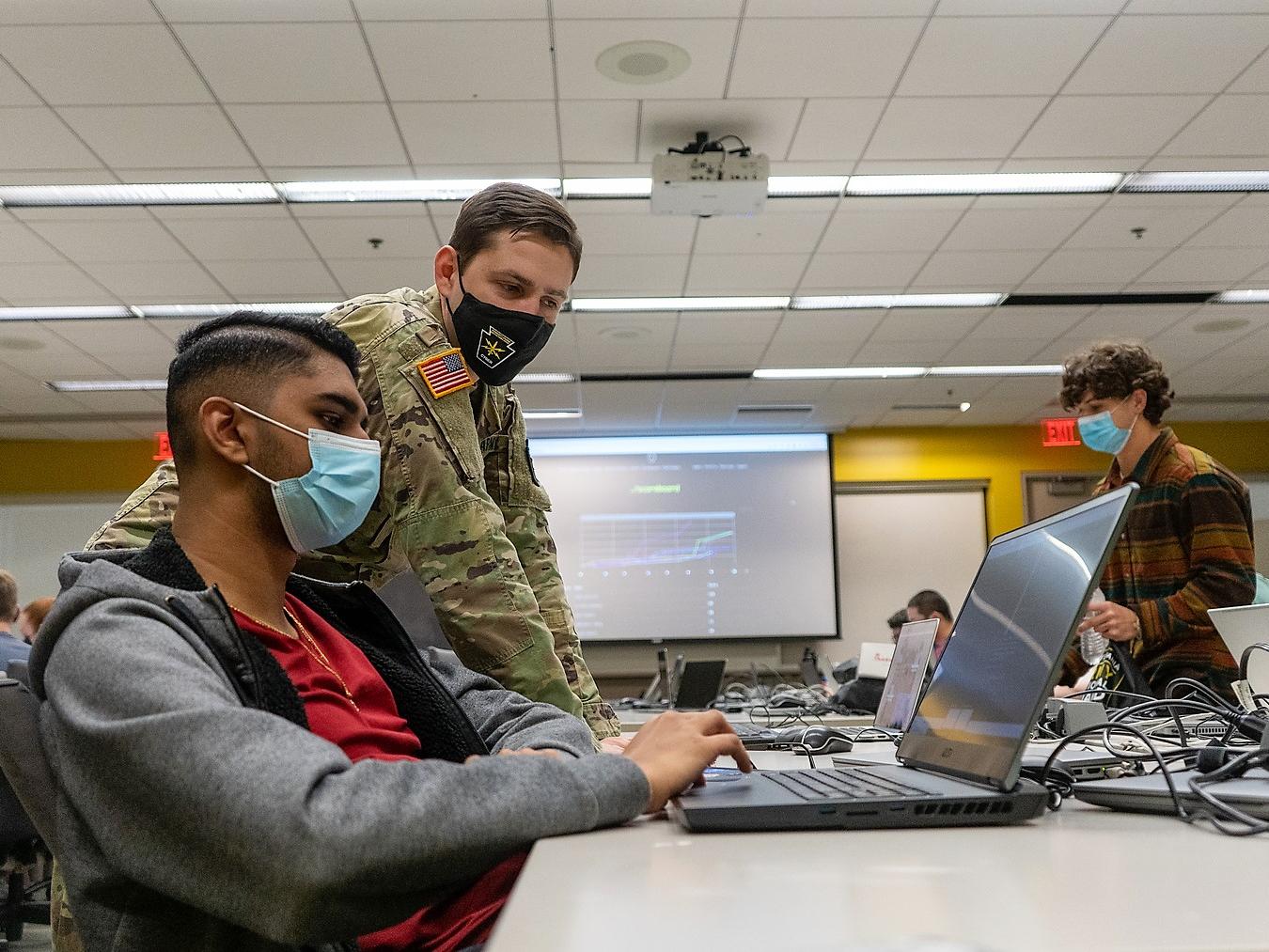 A PA National Guard member dressed in a a camoflauge uniform helps a student working on a laptop.