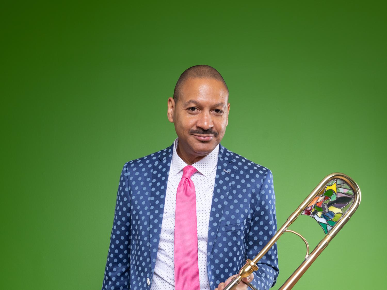 A man wears a suit and leans against a wall while he holds a trombone.