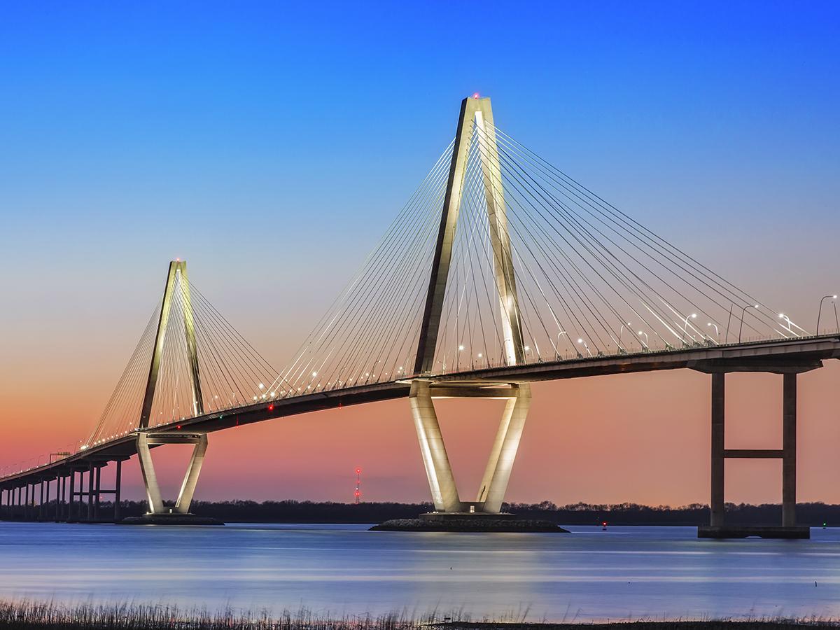 A suspension bridge over water is illuminated as the sun sets in the background. 