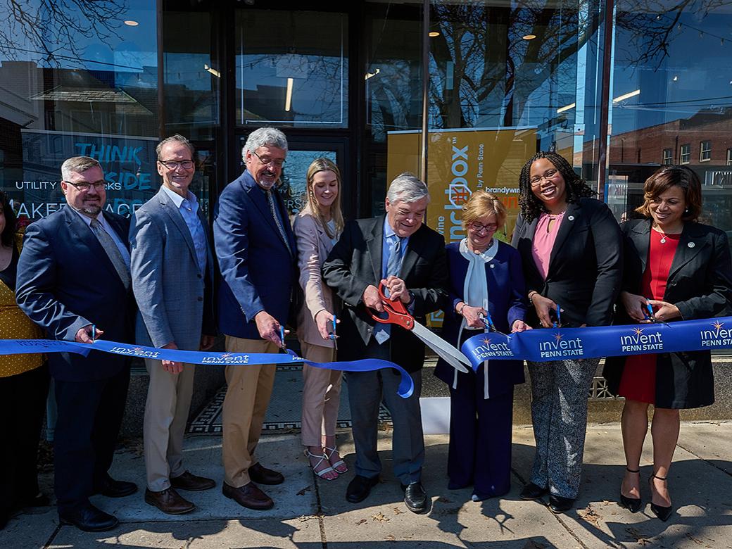 A group of 9 people cuts a long ribbon in front of a building.