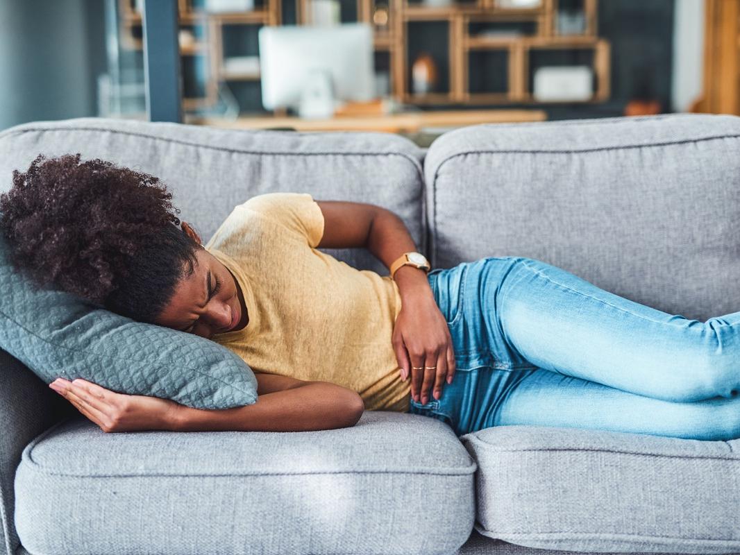 A young woman experiencing abdominal pain lies on the sofa at home.