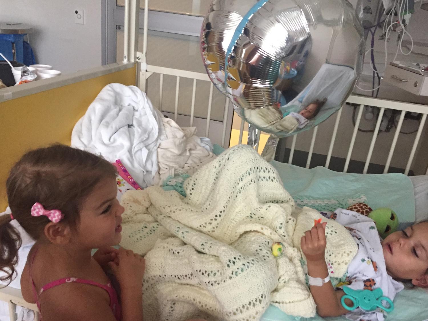 A young girl with pigtails and a ribbon in her hair visits another young girl in a hospital bed.