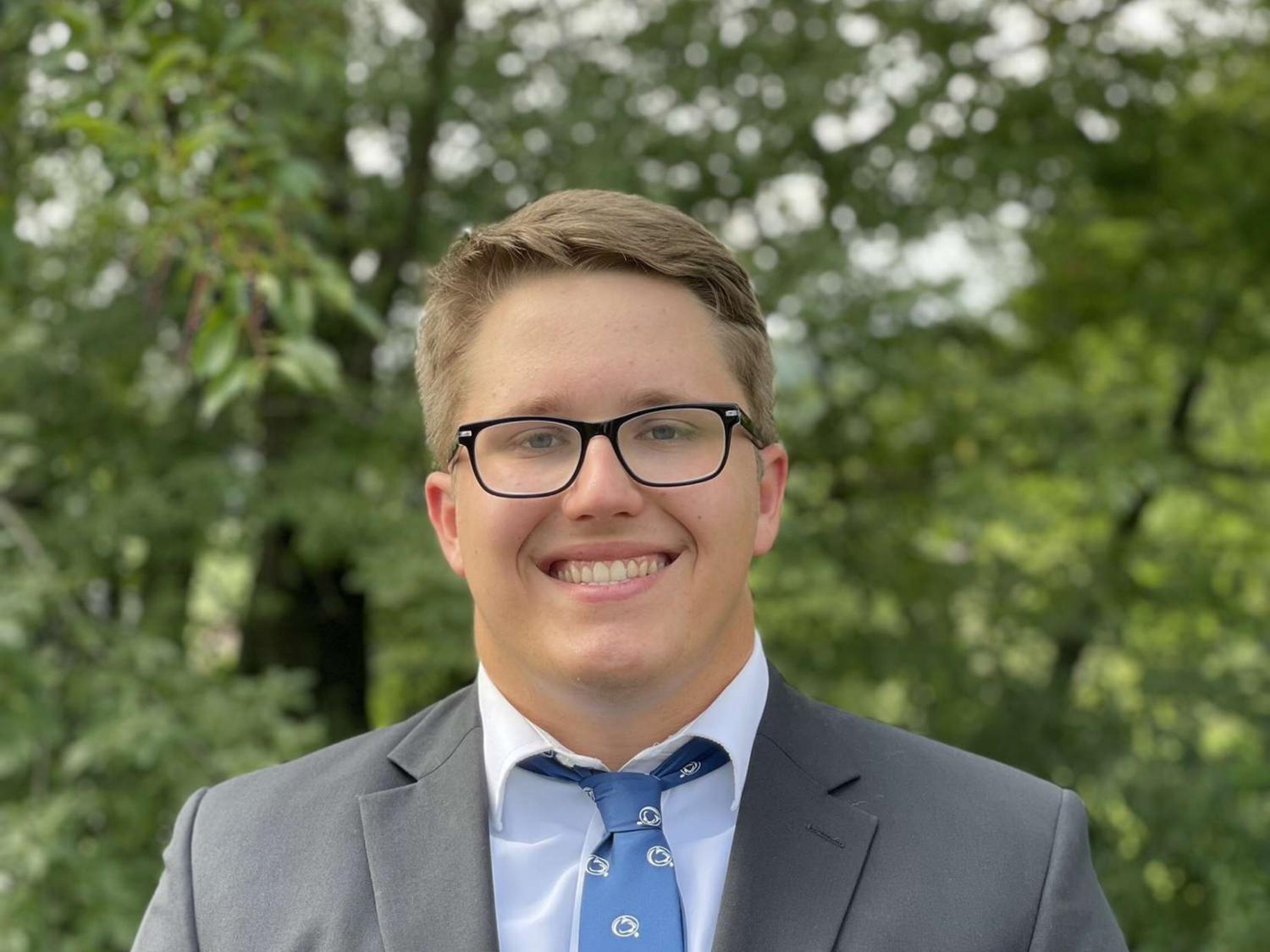 A male student smiling and wearing a suit and tie with trees in the background