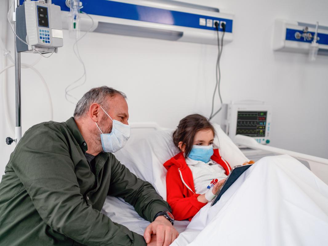 A young girl sits up in her hospital bed, playing on a tablet computer. She’s hooked up to medical equipment. A man seated at the side of the bed looks at the screen with her.