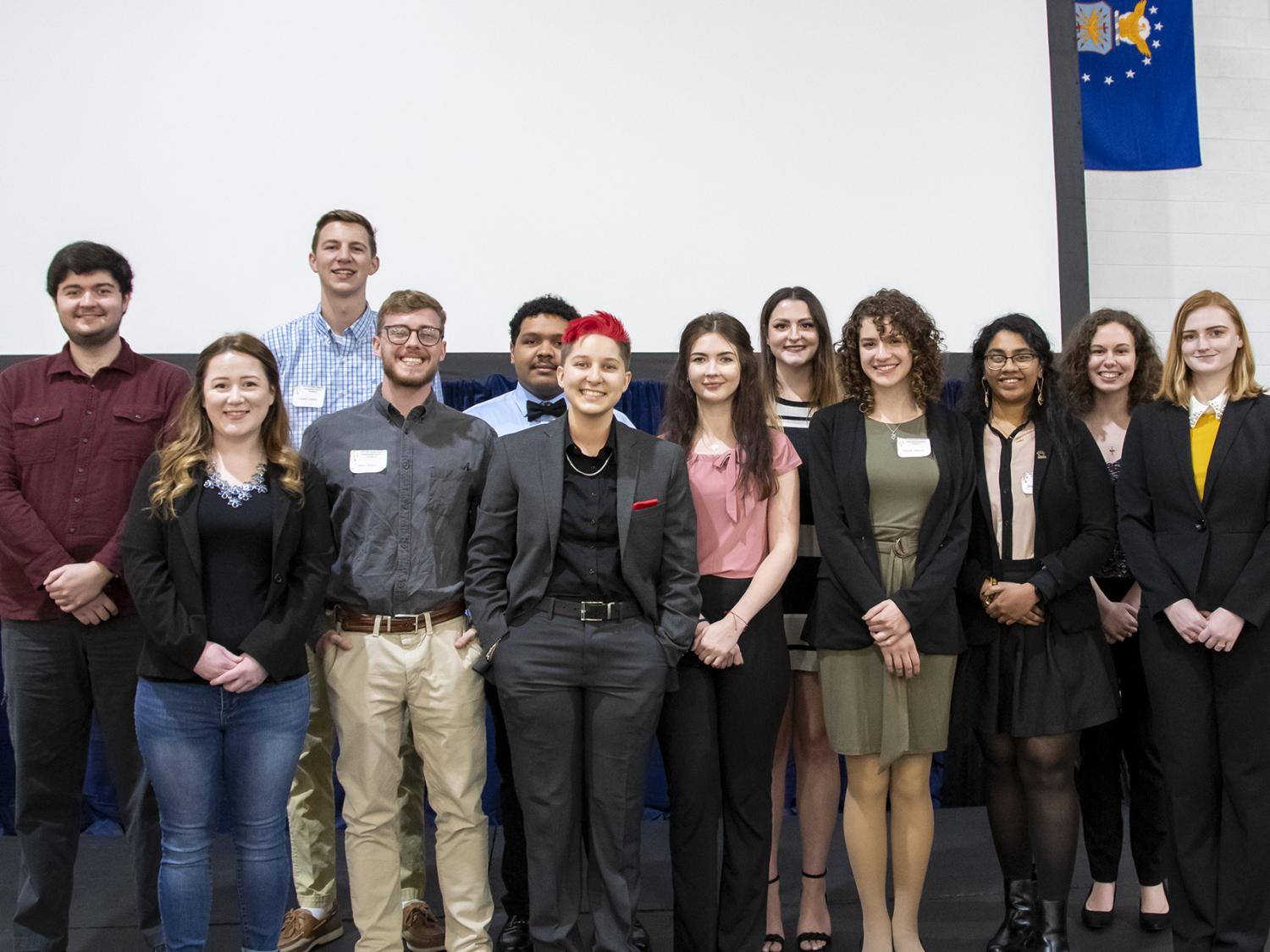 A dozen Penn State students pose for a photo on stage in front of a projector screen
