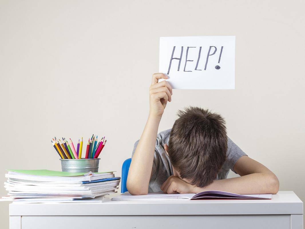Sad tired frustrated boy sitting at the table with many books and holding help sign.