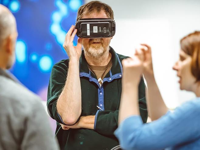 One person wears a virtual reality headset while two other people discuss the technology.