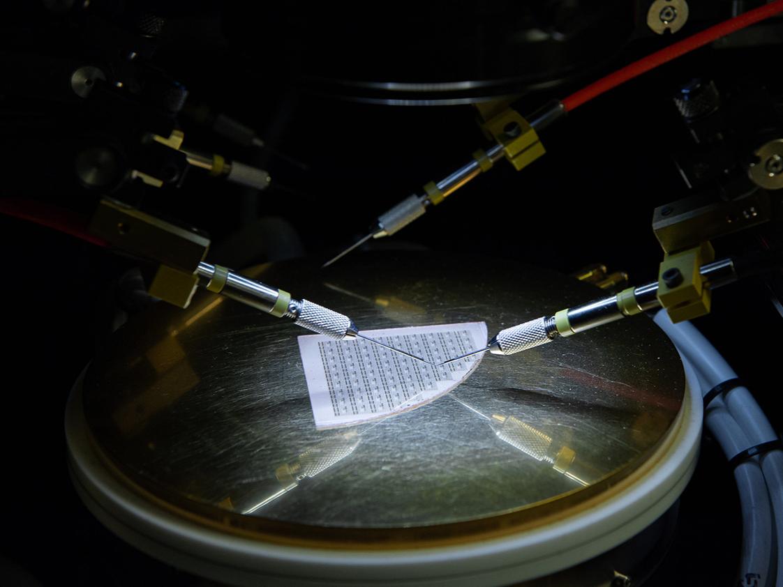 A white square with black lines sits on a metal circle, surrounded by three thin needles attached to larger machinery. 