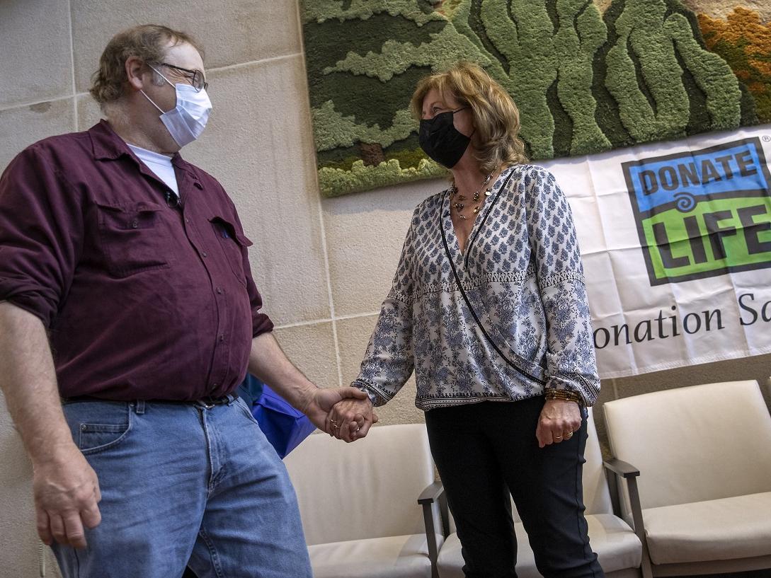 Dwayne Heller, who wears glasses and a face mask and is dressed in a chamois shirt and jeans, clasps hands with Maureen Stathes, who has shoulder length hair and wears a face mask, blouse and pants. A “Donate Life” banner is on the wall behind them.