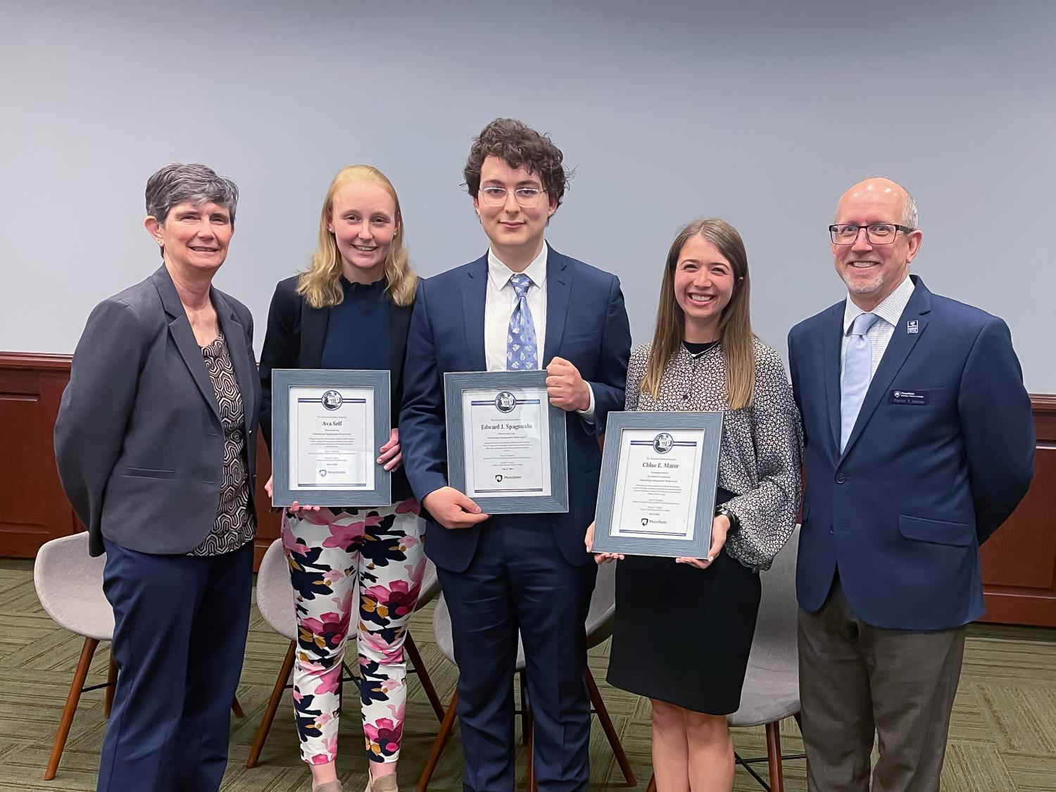 five people standing: two female students and male student in center holding framed certificates, female dean at far left and male dean at far right