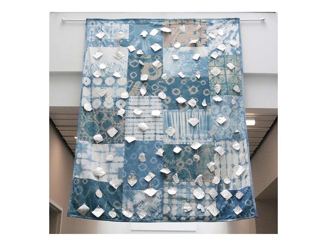 Indigo-colored quilt with medallions affixed to the front hangs in the hallway of a well-lit building.
