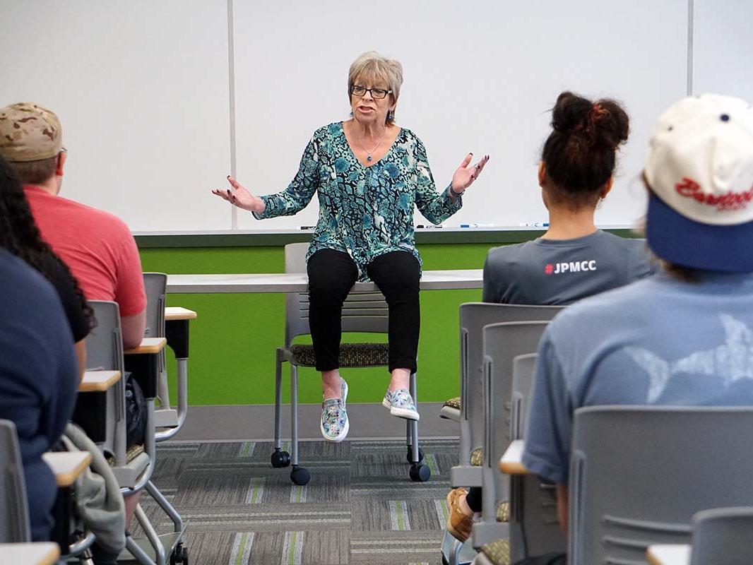 Woman seated on classroom table with rows of students on either side facing her.