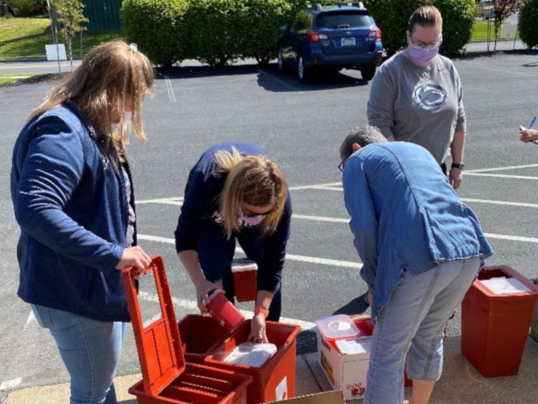 Four health care workers organize plastic containers full of syringes, which are placed at the edge of a parking lot. A car and bushes are in the background.