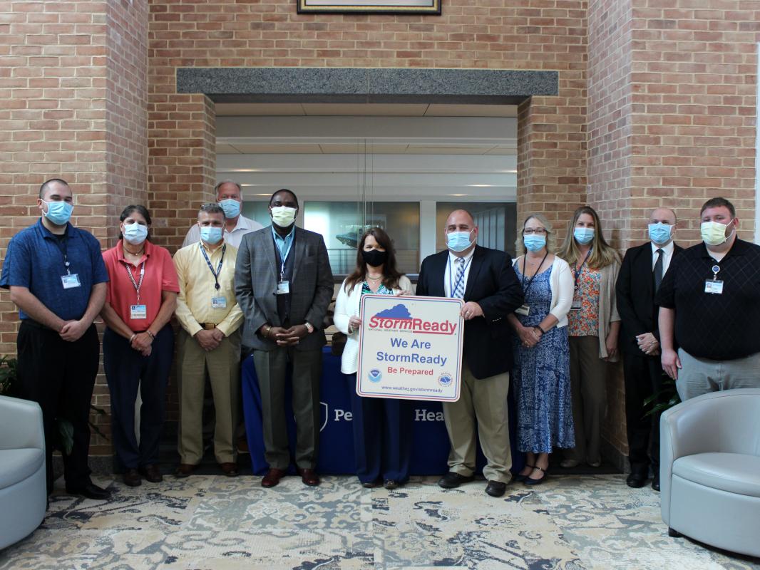 Eleven people stand in a row, posing with a certificate that reads “StormReady: We Are StormReady. Be Prepared.” A brick wall with an opening in the middle is in the immediate background.