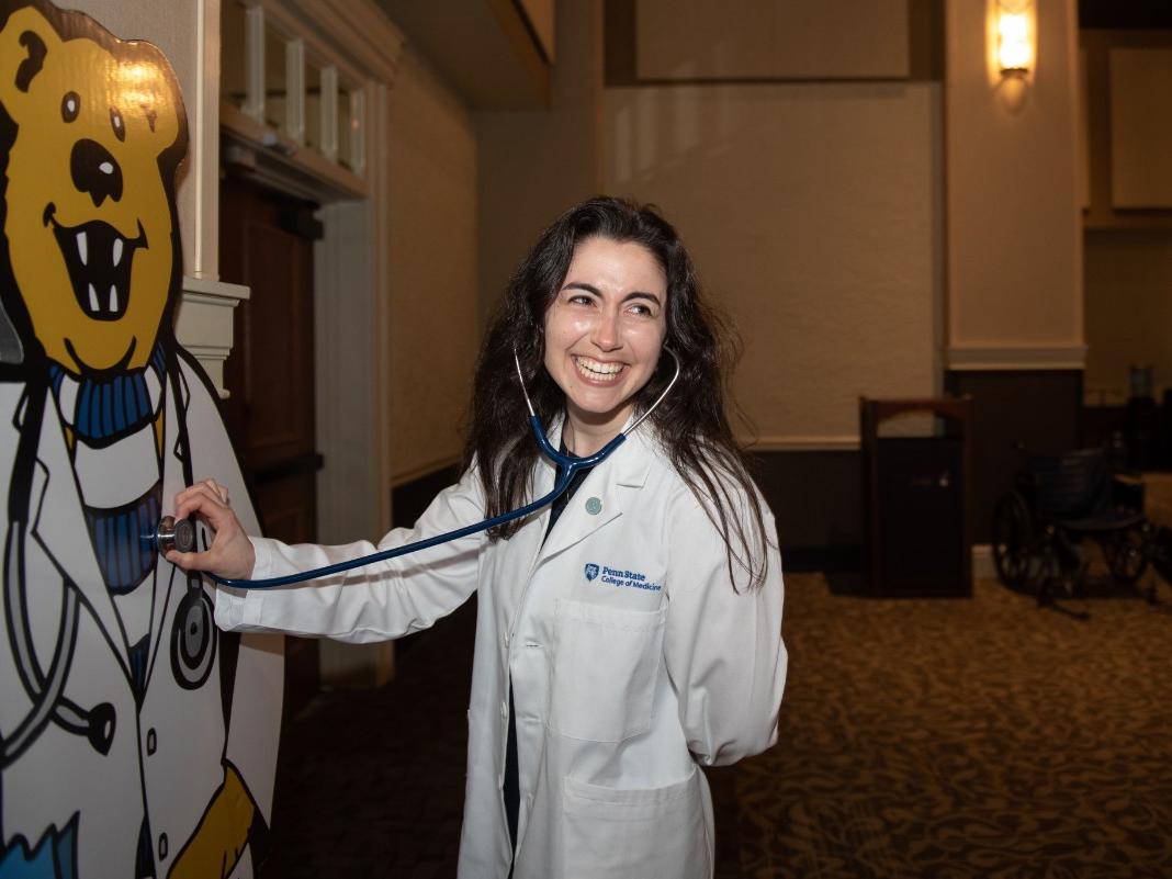 A woman wearing a physician assistant coat holds her stethoscope up to a Nittany Lion cardboard cutout that is also wearing a physician assistant coat.