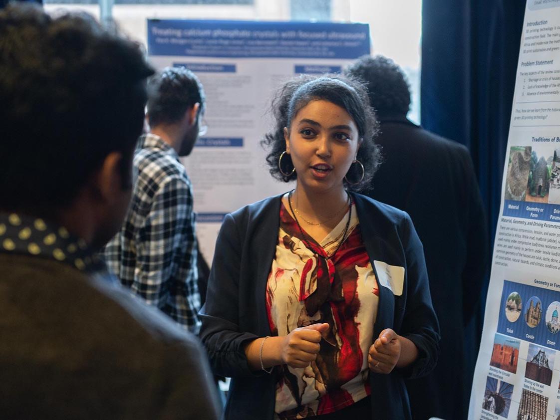 A woman stands in front of a large poster and explains her research to a person in the foreground.
