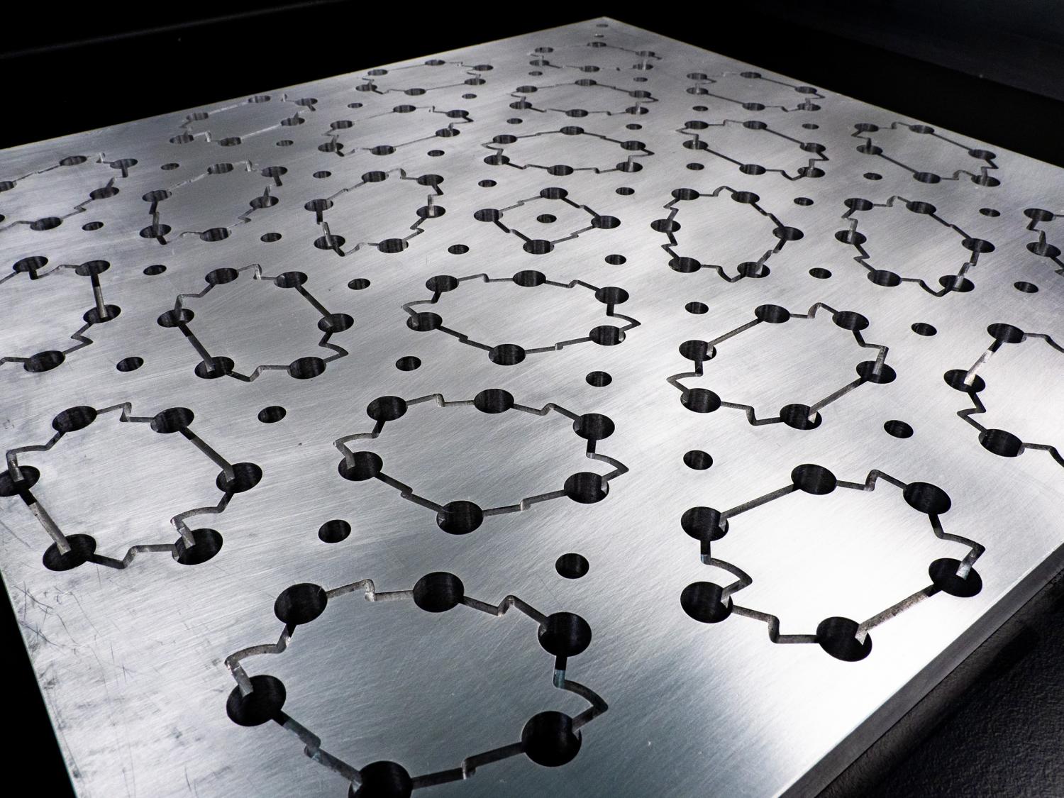 A metal surface with small, dark holes spaced in a rough hexagon pattern. In a regular cadence, jagged etched lines connect the holes to form the individual hexagons, separated by unconnected smaller holes. 
