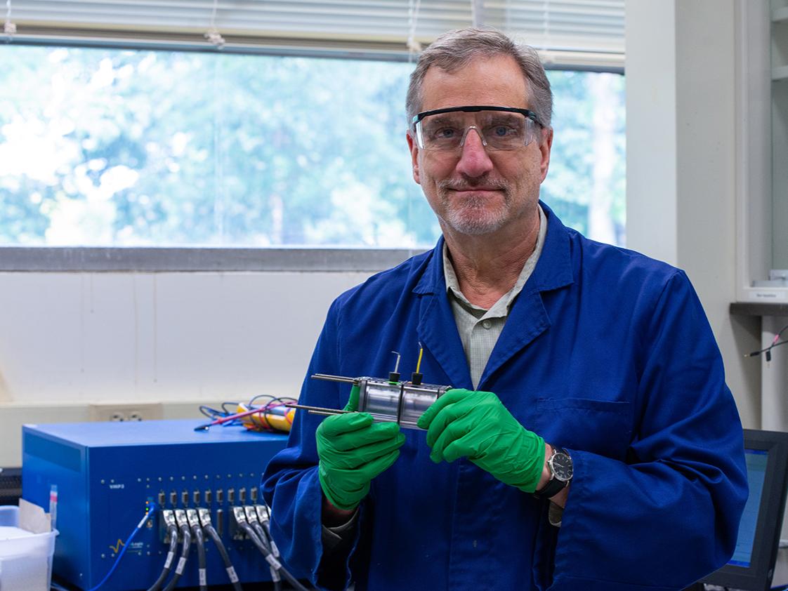 A man in a blue lab coat and protective glasses holds a small electrochemical reactor.