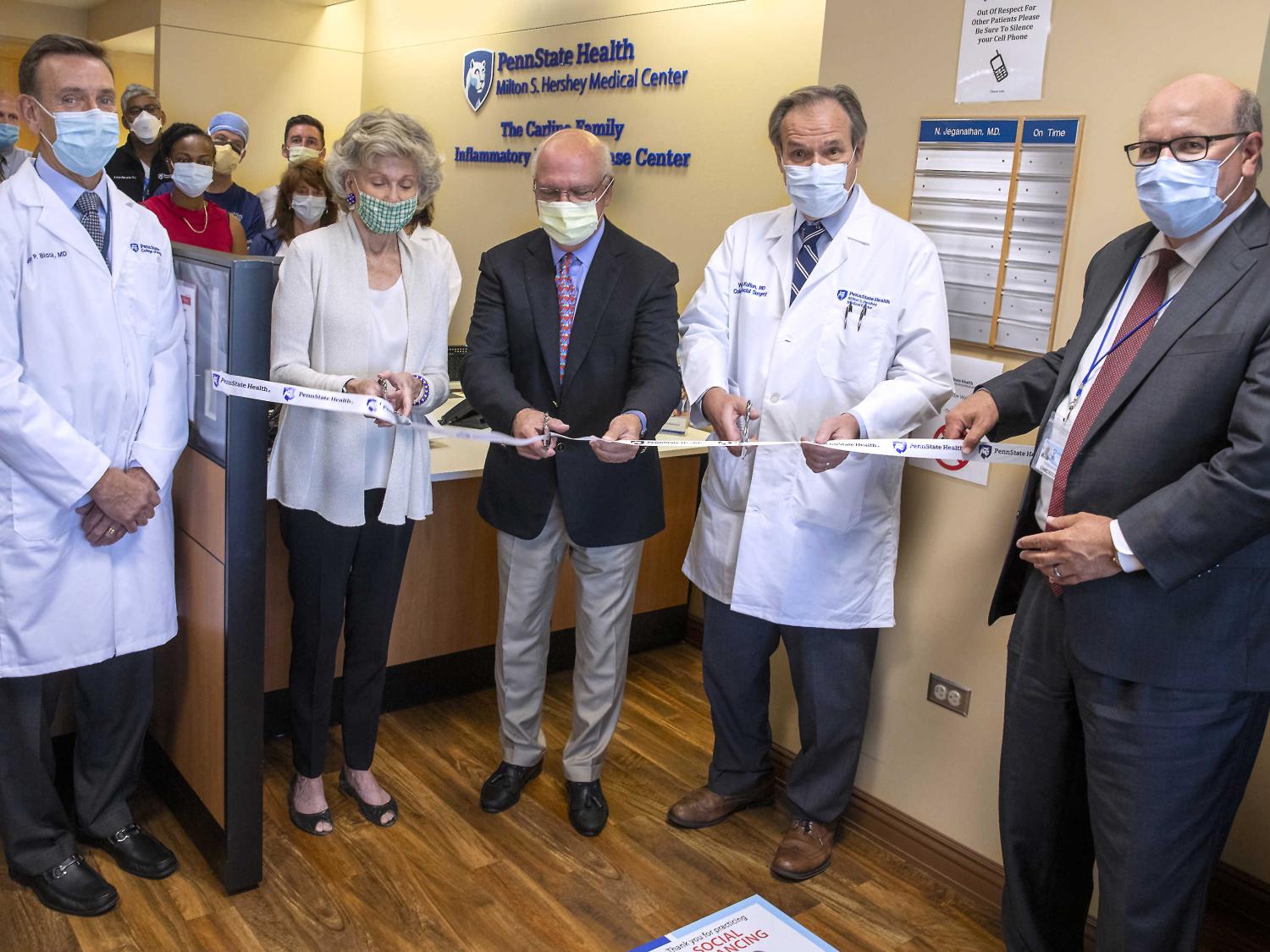Five people in the foreground pose for a photo as they prepare to cut a ceremonial ribbon. A small group of people looks on in the background.