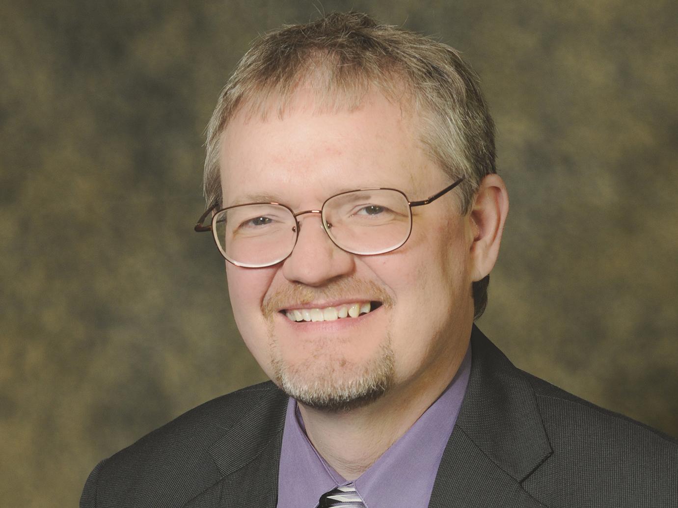 A headshot image of Daniel Christopher wearing a gray suit and purple shirt. 