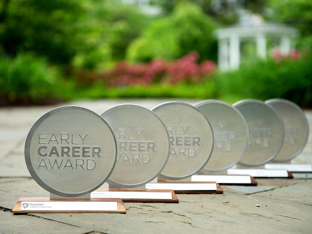 Six awards are lined up on stone, with greenery in the background. Each Award is a silver circle erected on a wood stand.