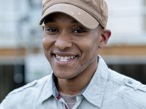 A man smiles at the camera wearing a brown hat and a blue collared shirt. 