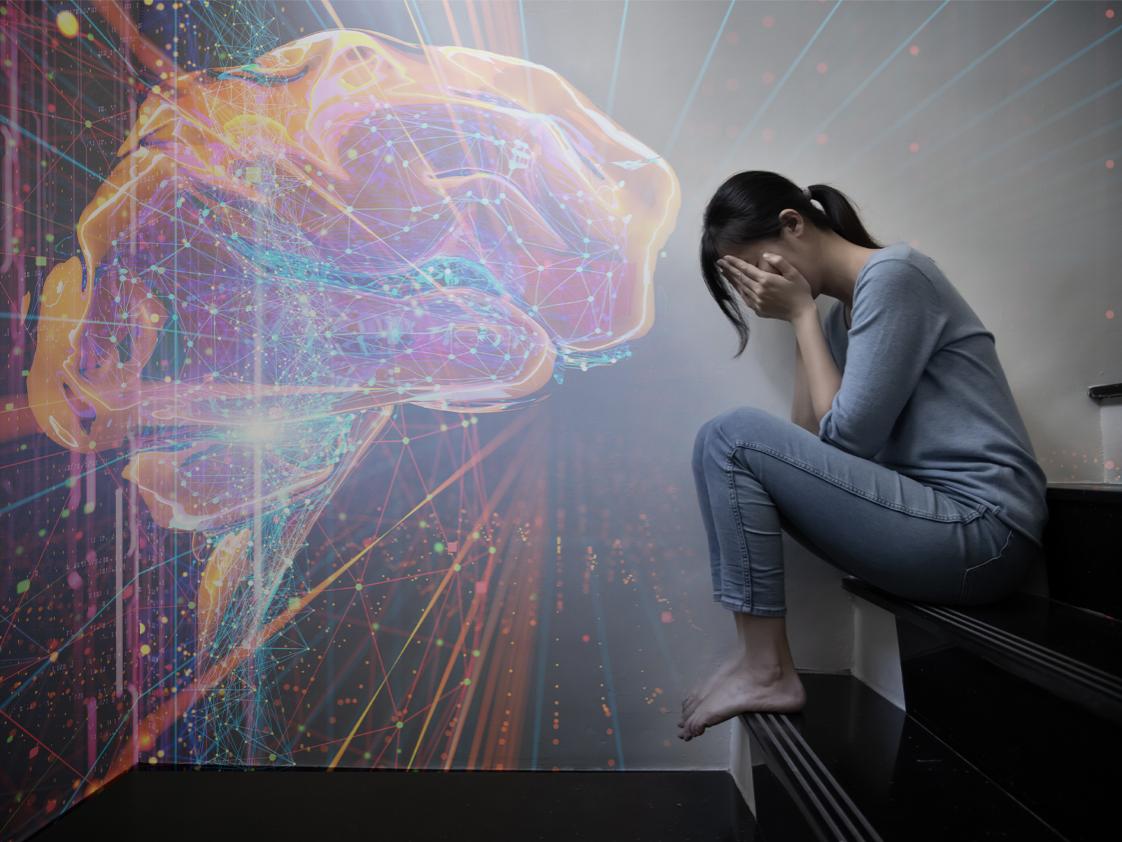 On the right side of the image, a person sits on a step with her face in her hands. A digital rendering of a brain is projected on the left side of the image. 