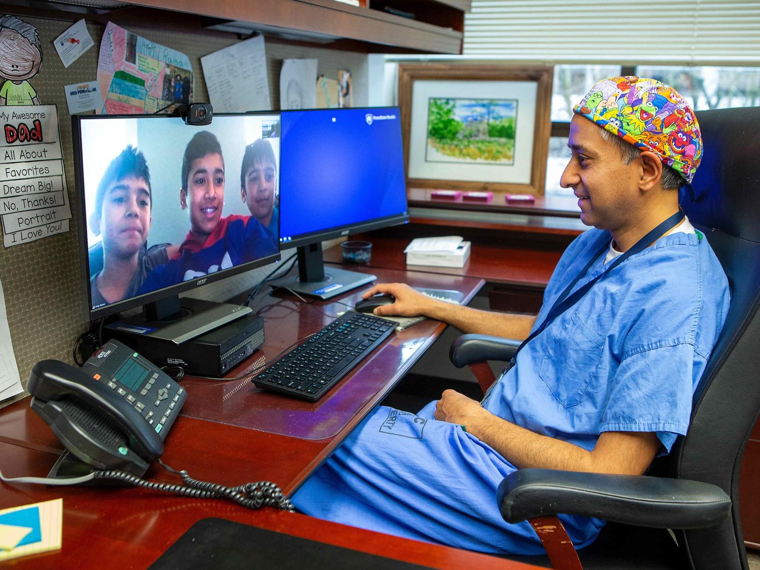 A doctor in scrubs smiles at images of three boys on his computer screen.