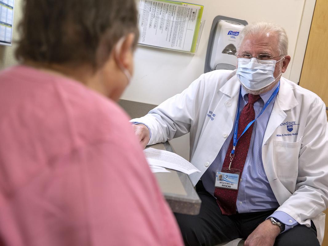 Dr. Witold Rybka, who wears a face mask, glasses and a white coat, sits in an exam room opposite a patient, who is seen from behind. A hand sanitizer dispenser and rack of papers are mounted on the wall behind him.