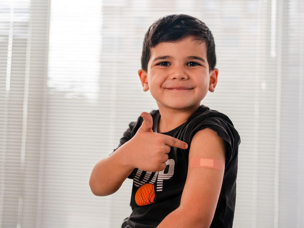 A vaccinated child points to an adhesive bandage on his arm, with his shirt sleeve rolled up.