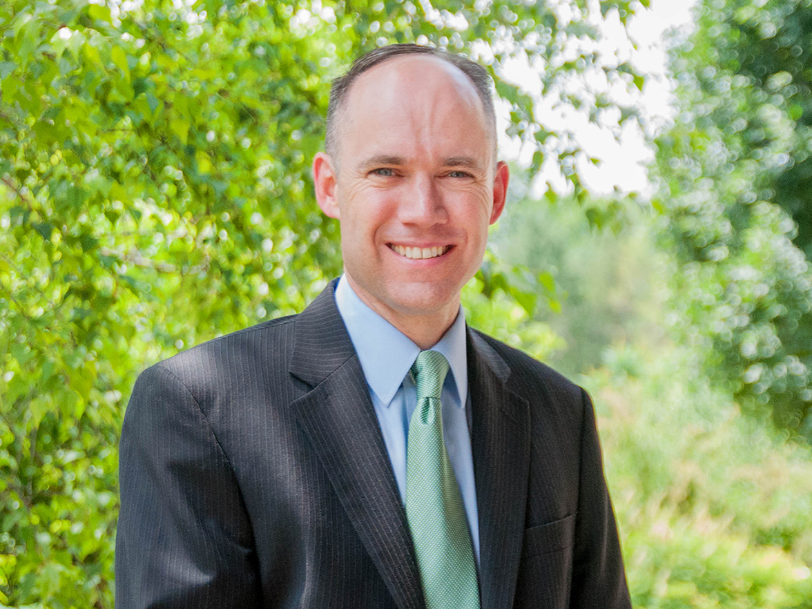 Man wearing a suit and smiling in an outdoor setting with trees behind him