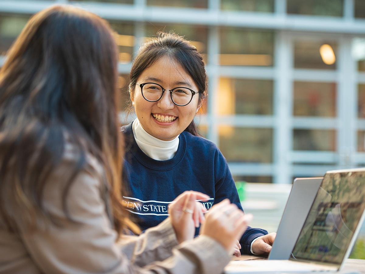 A photo of two female students sitting with their laptops outside of the Business Building.