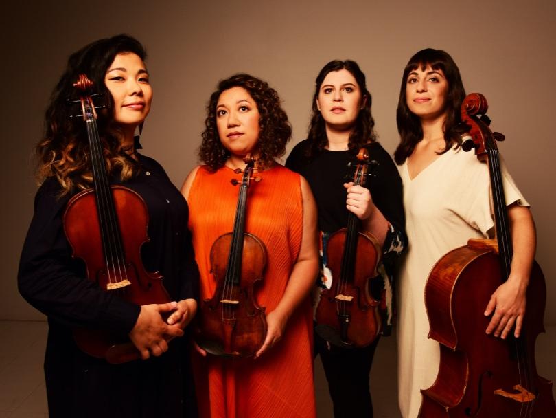 Four woman musicians stand shoulder to shoulder and hold their stringed instruments.