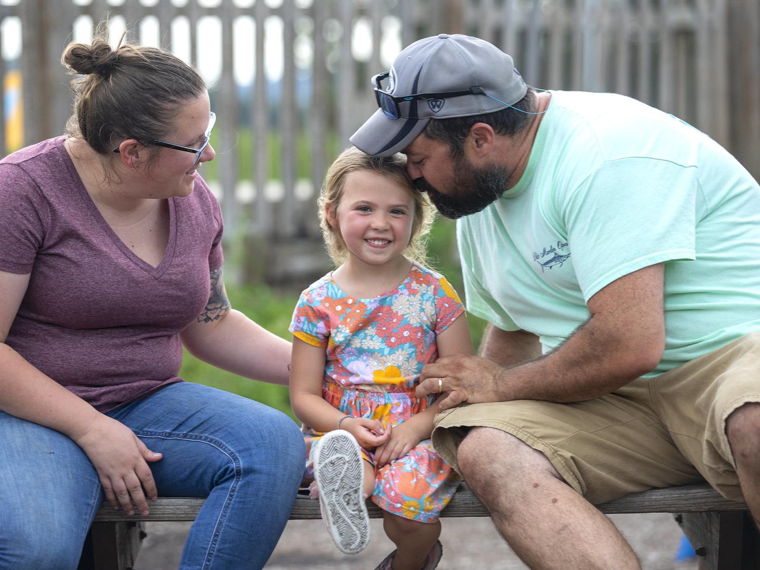 A man and a woman to the left and right of a little girl in a dress on a park bench. The little girl smiles as the man leans down and nuzzles the side of her head.