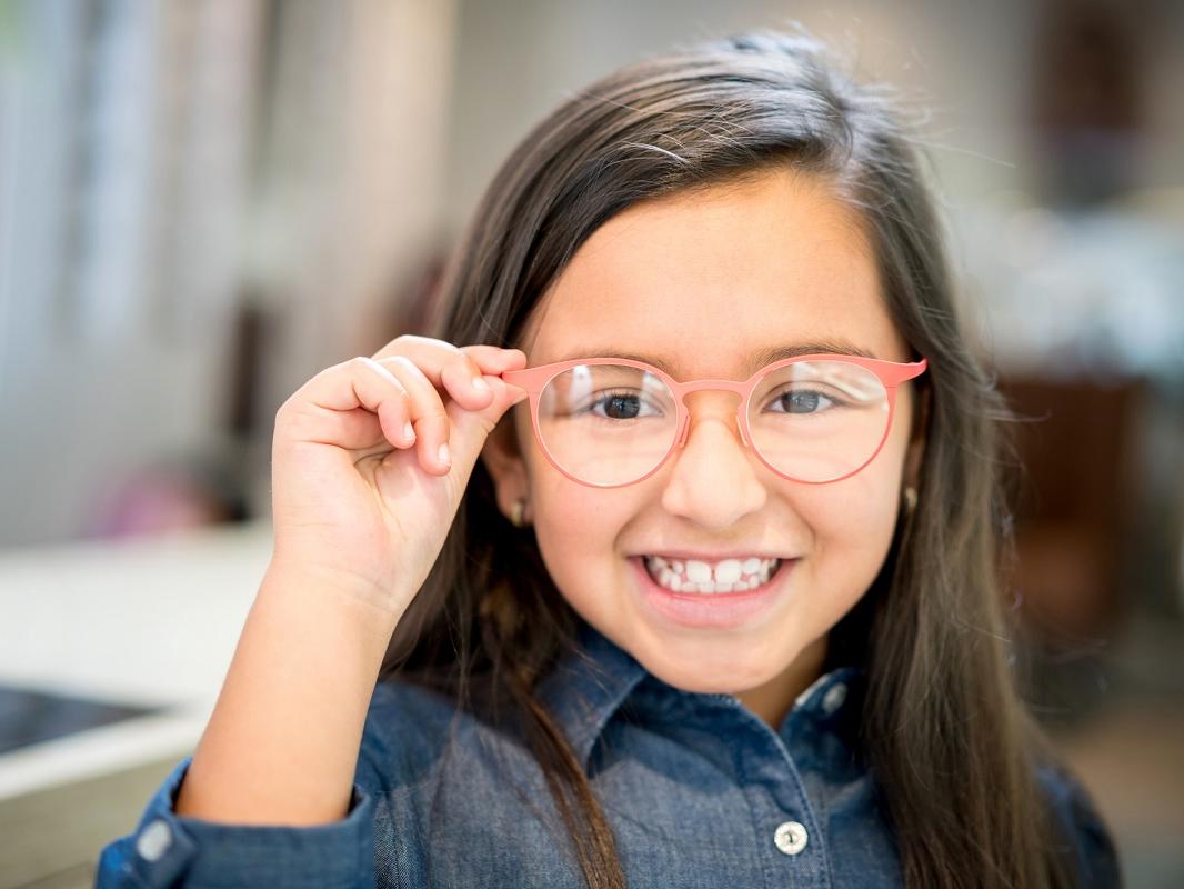 A young girl tries on glasses, smiling toward the camera.