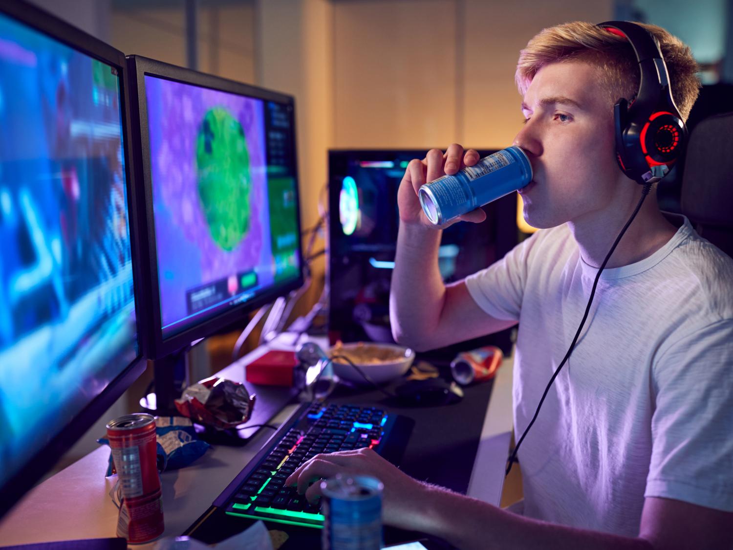 young man sits at computer surrounded by energy drink cans and chip bag wrappers. He drinks from a can while typing with his other hand