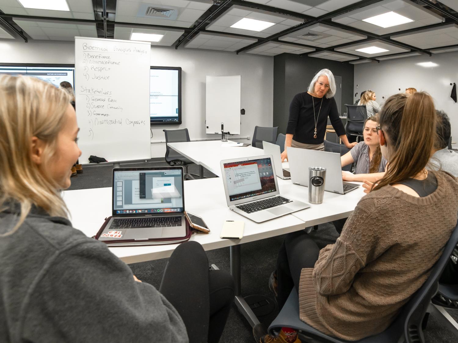 An instructor stands by a group of students who are seated with laptops in a classroom 