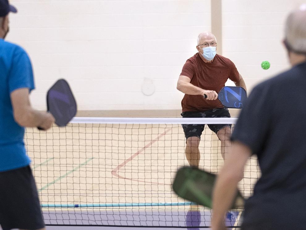 A man in a surgical mask charges a net brandishing a pickleball racquet in a gym. Two other men are seen from behind in the foreground.