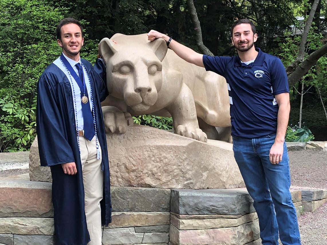Male graduate in cap and gown and another male student standing next to Nittany Lion statue.