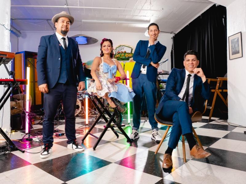Four people wearing dapper formal clothing pose in a room next to keyboards.