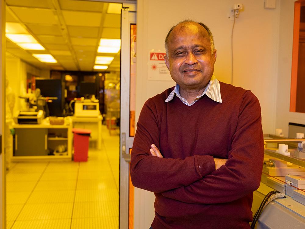 A person stands and smiles at the camera. Lab equipment is visible in the background. 