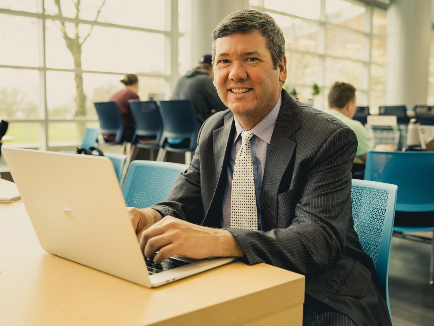 A photo of Terrence Guay sitting with his laptop in the Business Building Atrium.