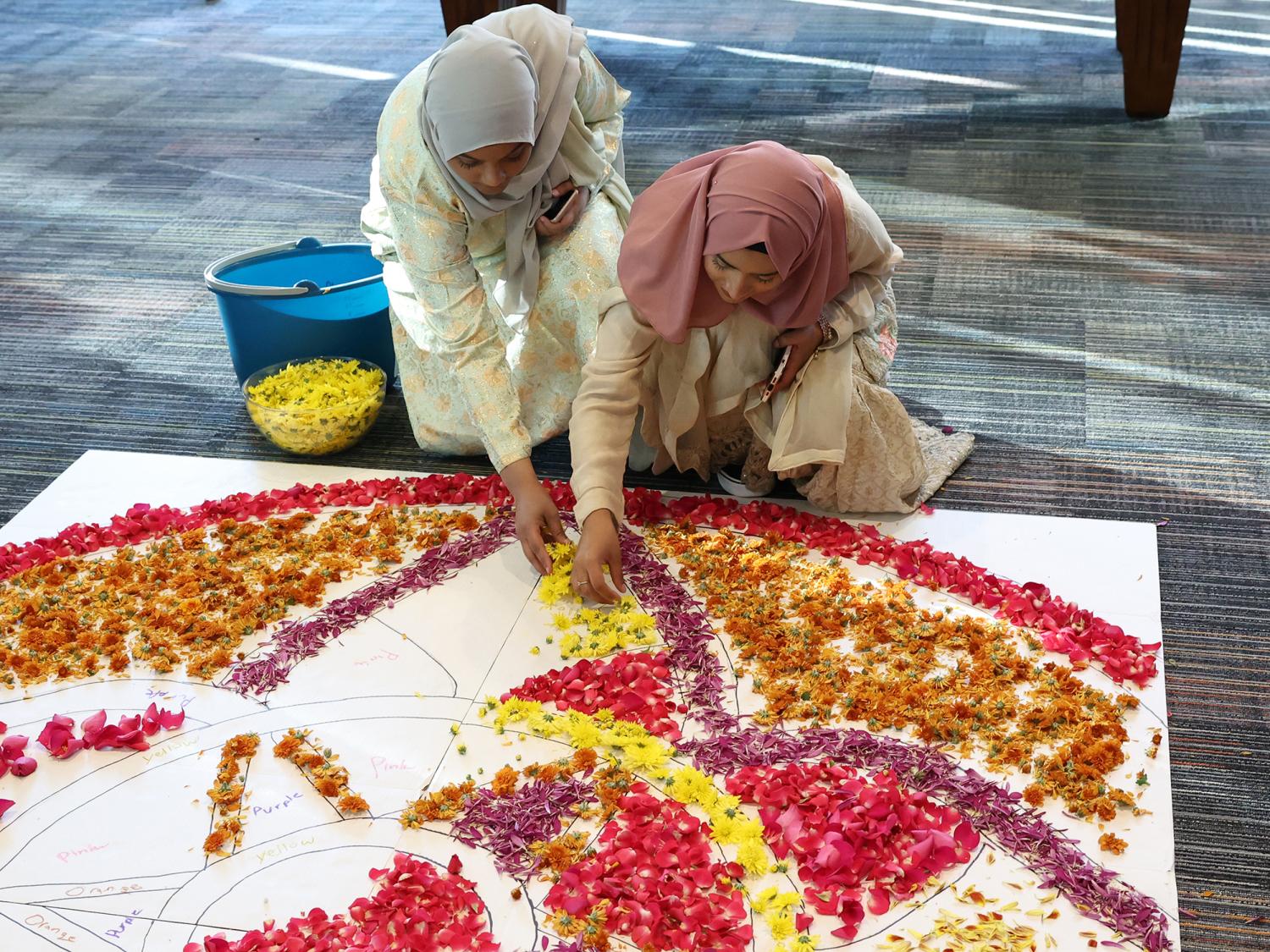 Two female students place flower petals onto the floor to create a rangoli.