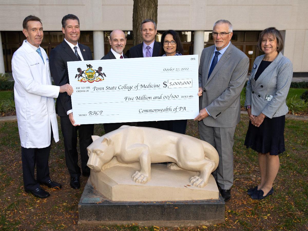 Seven people pose for a photo behind the Nittany Lion statue at Penn State College of Medicine. They are holding a commemorative check for $5 million.