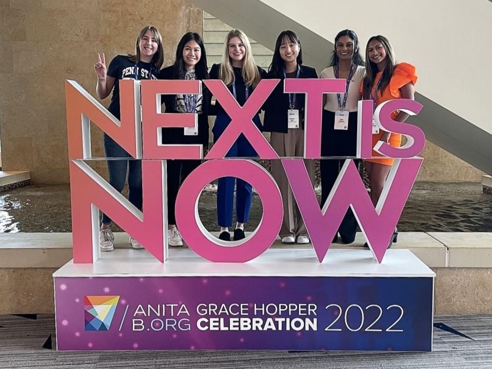 Six IST students behind a sign that reads Next is Now