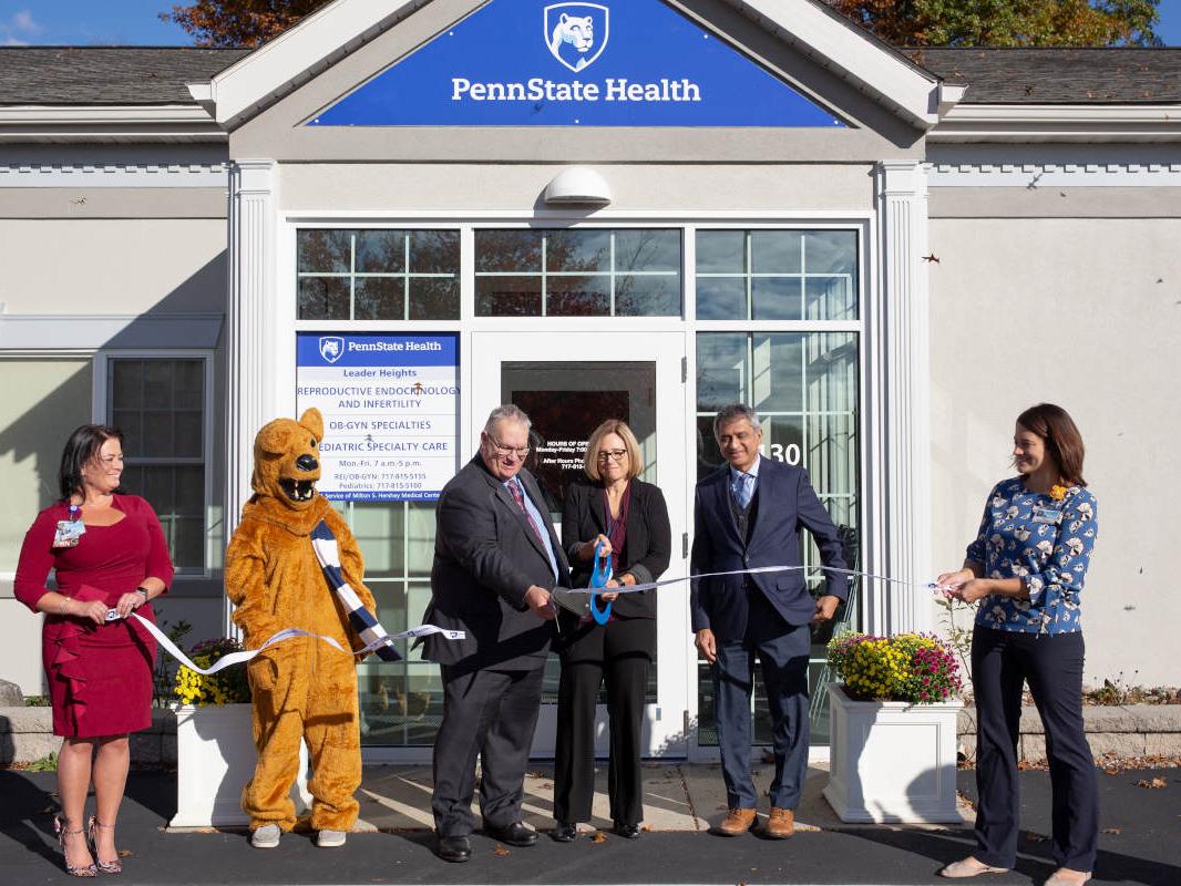 Six people, including one person dressed as the Nittany Lion, stand in a row outside the front door of a building. They pose for a photo as they all hold onto a ribbon. The Penn State Health logo is on the entranceway, above them.