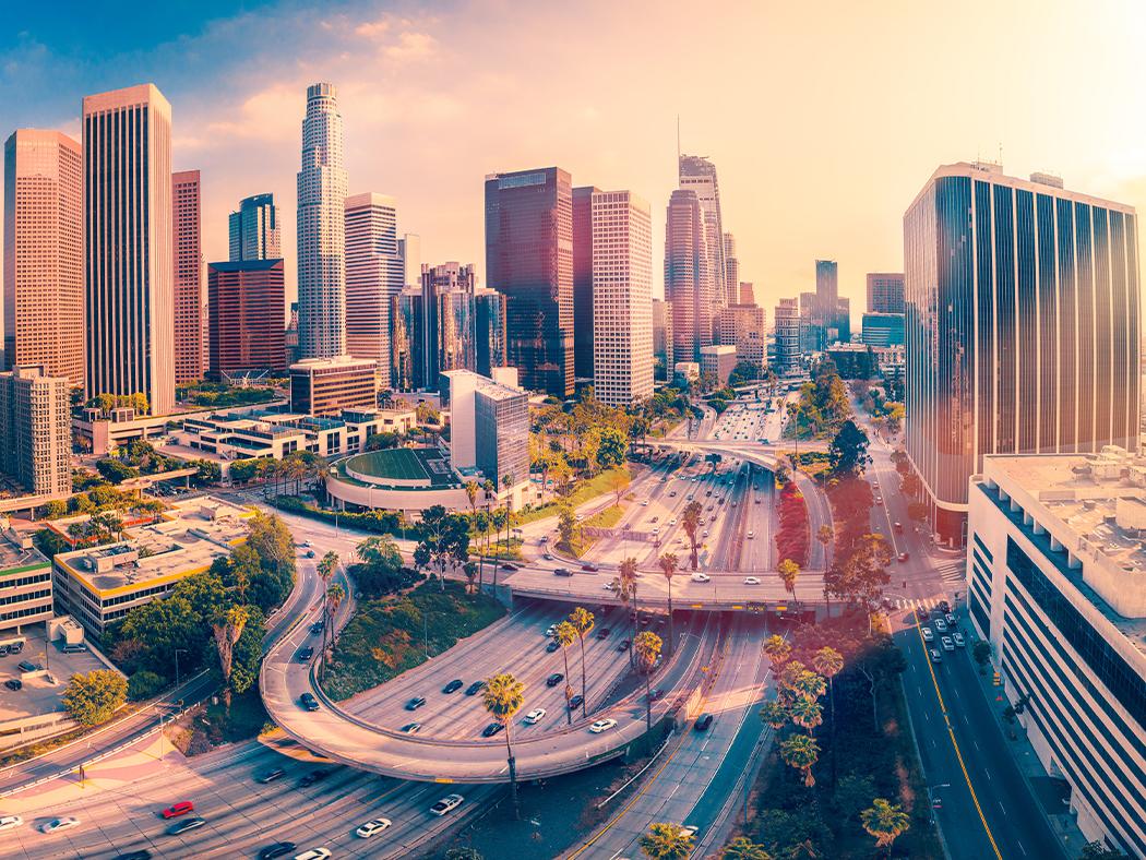 An image of the LA Skyline with the sun hitting it from behind with an overpass in the foreground. 